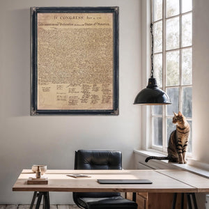 a framed document titled "IN CONGRESS, July 4, 1776" hanging on a wall above a wooden desk with a cat sitting on the windowsill.