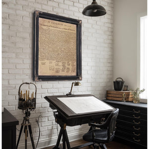 a workspace with a desk, a lamp, and a framed document hanging on the wall. The desk has a drawing pad and a lamp on it, creating a functional and organized environment.