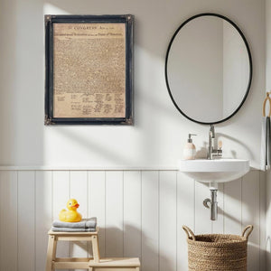 a bathroom with a framed document hanging on the wall, a white sink, a round mirror, a wooden stool, and a basket.