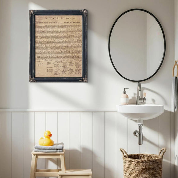 a bathroom with a framed document hanging on the wall, a white sink, a round mirror, a wooden stool, and a basket.